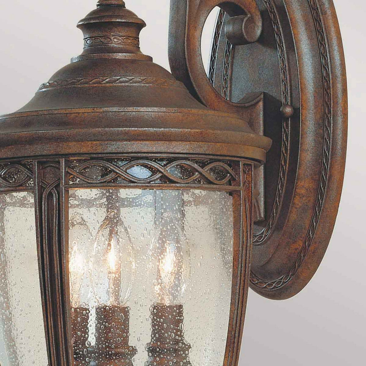 Close-up of the English Bridle 3 Light Outdoor Medium Wall Lantern - Bronze, showcasing its vintage-style antique bronze finish and clear seeded glass shade. The lantern features three visible candle-like light bulbs inside.