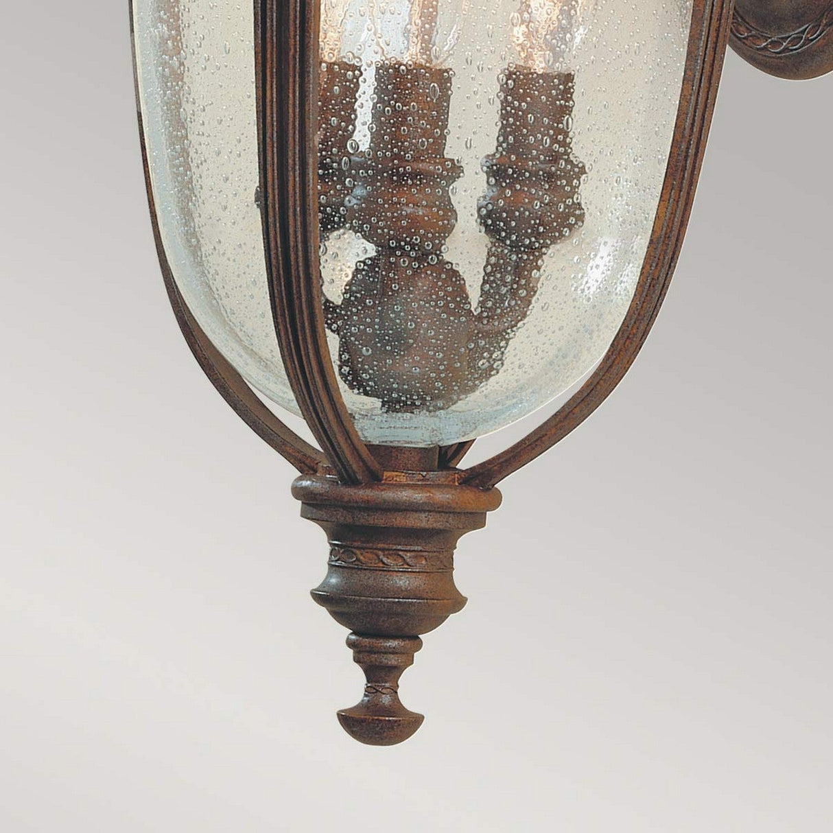 The English Bridle 3 Light Outdoor Medium Wall Lantern in Bronze displays a vintage aesthetic with its clear seeded glass and metal details. This fixture boasts an antique charm through its bronze lantern design and multiple candle-like bulbs, showcased against a light gray backdrop.