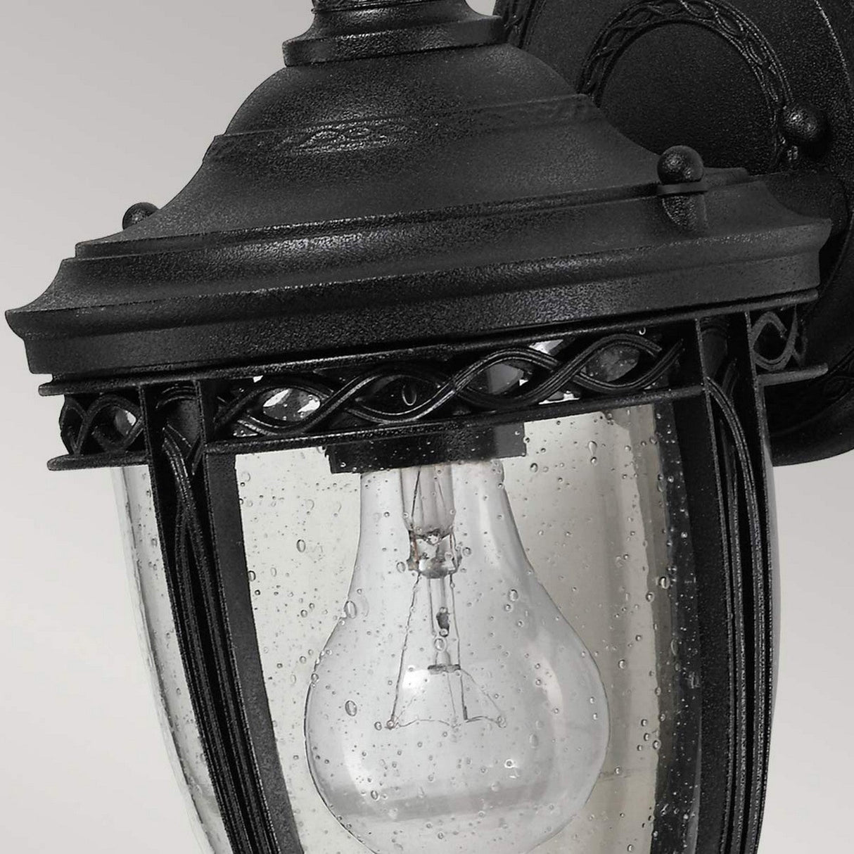 A close-up view of the English Bridle 1 Light Outdoor Small Wall Lantern in black finish, featuring a clear glass cover. The lantern showcases a visible light bulb and is adorned with decorative patterns, with droplets on its surface.