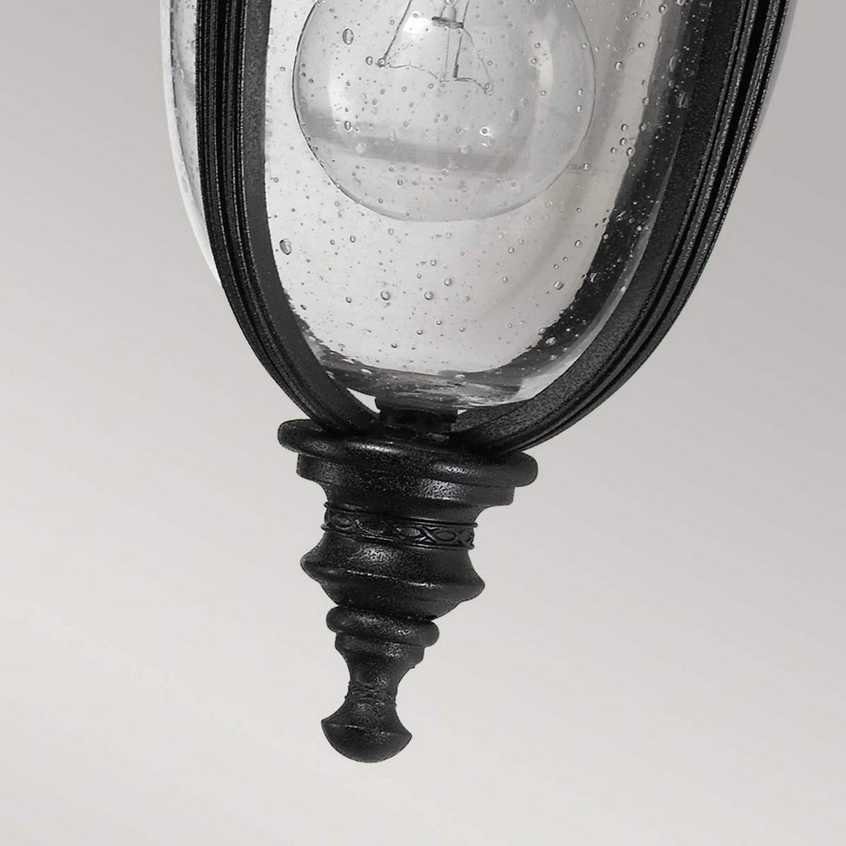 A close-up of the English Bridle 1 Light Outdoor Small Wall Lantern in black reveals its elegant design. The lantern features clear textured glass that showcases a visible bulb and includes an ornamental finial at the bottom, all beautifully set against a neutral gray background that enhances its sophisticated charm.
