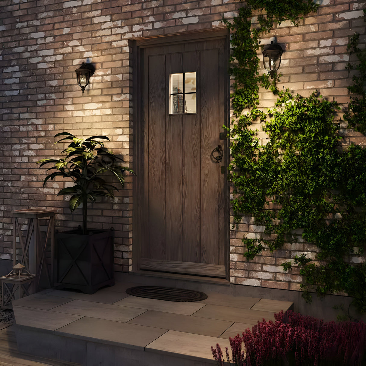 A rustic wooden door with a small window is set in a brick wall, surrounded by decorative ivy and two English Bridle 1 Light Outdoor Small Wall Lanterns in a sleek black finish. A potted plant sits on the left, complementing the stone porch's circular doormat and vibrant flowers.