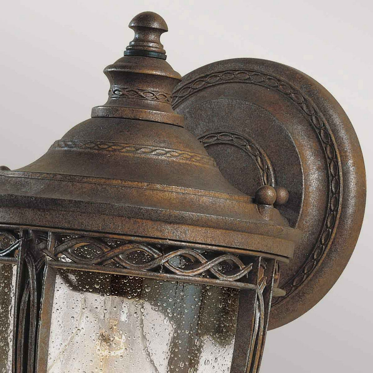 Close-up of the English Bridle 1 Light Outdoor Small Wall Lantern in a British bronze finish. The glass displays a textured raindrop pattern, complemented by decorative metalwork with an ornate design.