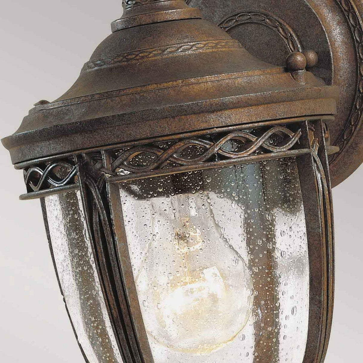 Close-up of the English Bridle 1 Light Outdoor Small Wall Lantern in a British bronze finish. The glass panels are adorned with a textured, water-drop design, and the filament bulb inside emits a warm glow. This outdoor wall lantern is elegantly mounted on a curved metal bracket.