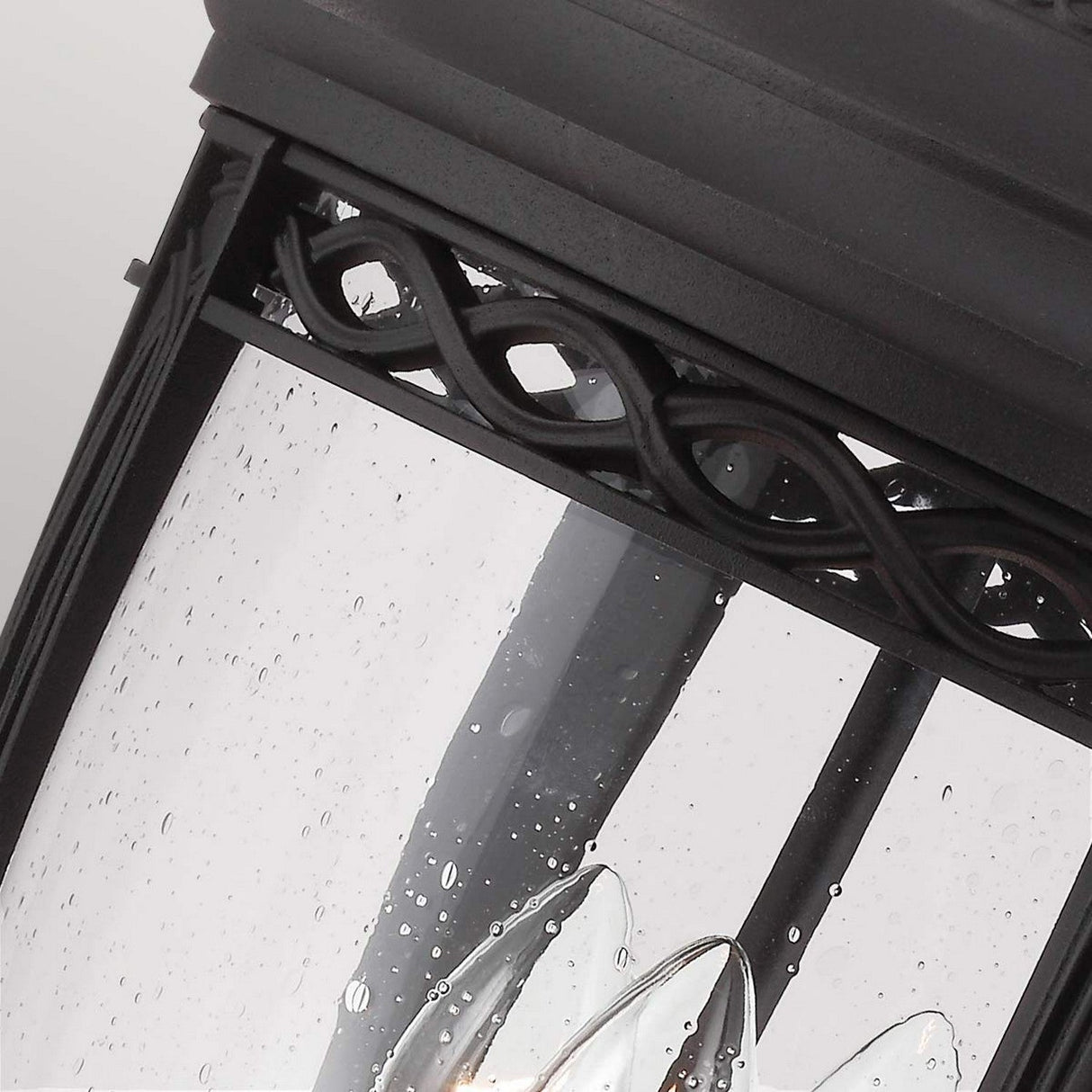 A close-up of the English Bridle 3 Light Outdoor Large Pedestal in black showcases its decorative loop design near the top, with raindrops adorning the glass panes. Perfect for outdoor lighting, it rests elegantly as a pedestal light, where a candle-shaped bulb emits warmth through its sleek black finish.