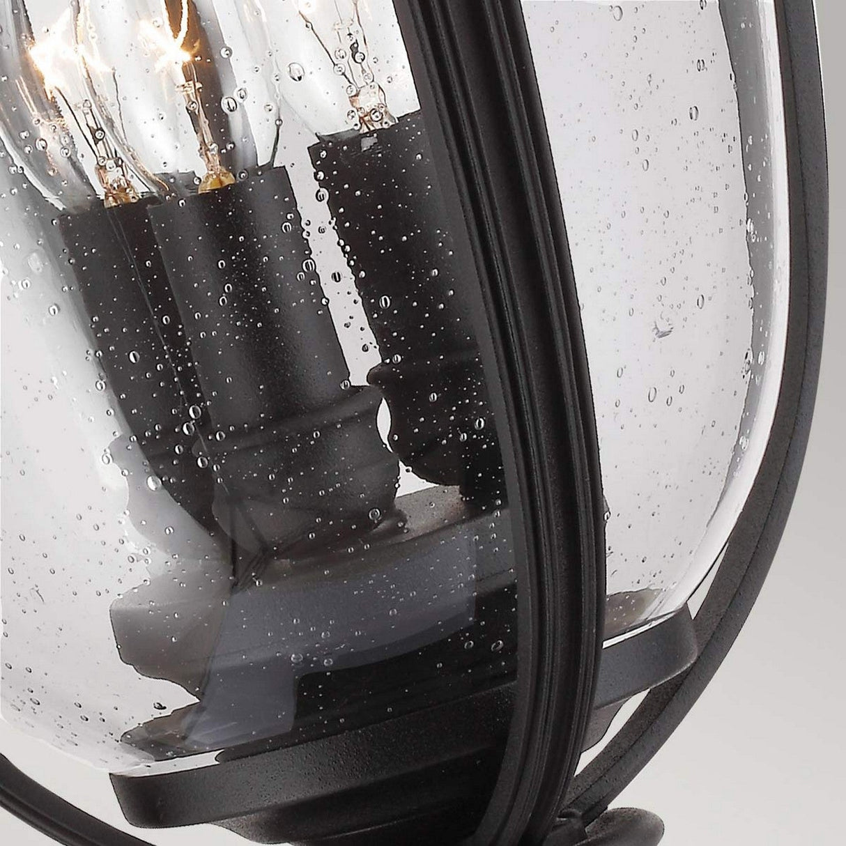 Close-up of the English Bridle 3 Light Outdoor Large Pedestal - Black with raindrops on its clear glass surface. The sleek black finish frame encloses lit bulbs, casting a cozy, warm glow against the droplets, capturing the essence of refined outdoor lighting in a rainy scene.