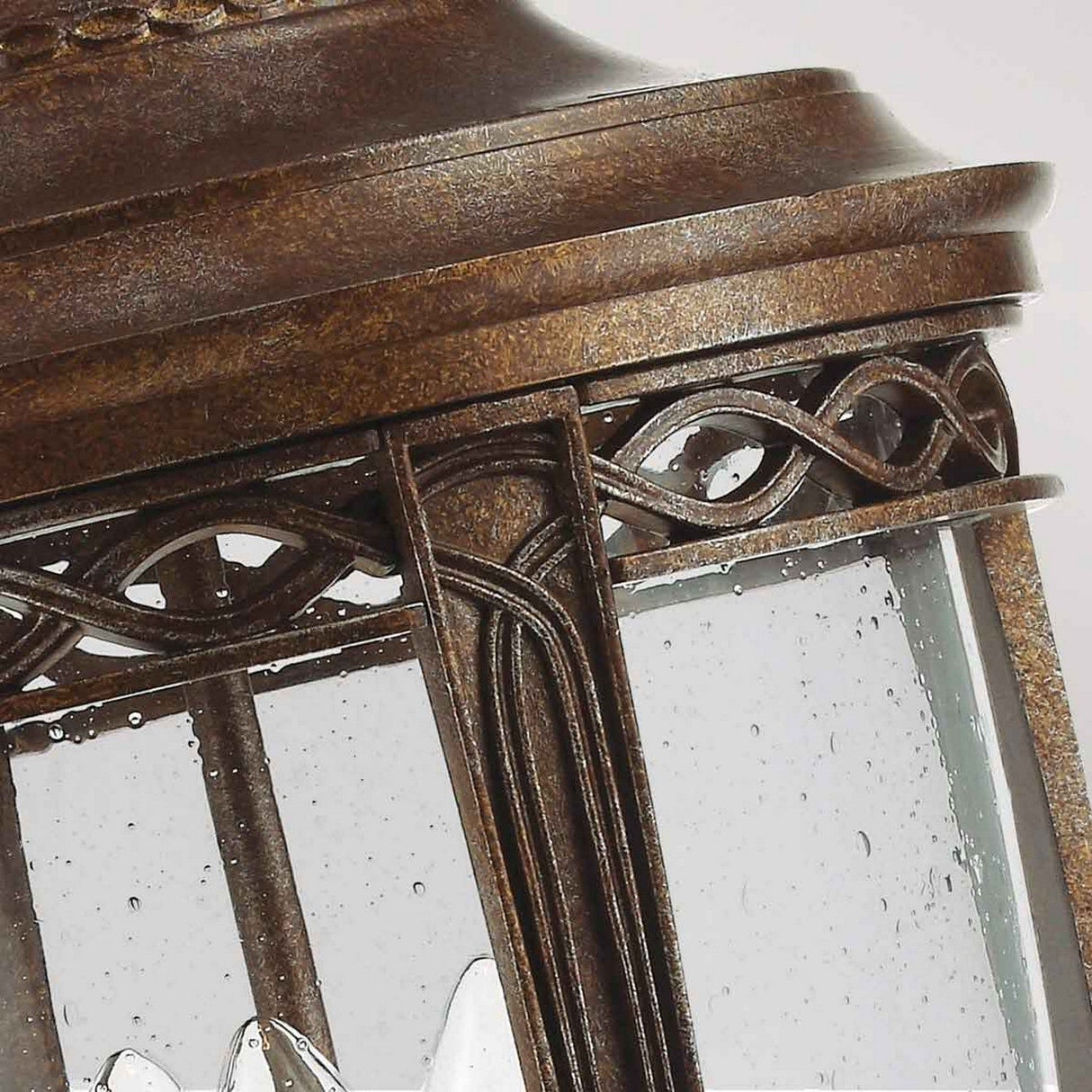 A close-up of the English Bridle 3 Light Outdoor Large Pedestal in bronze highlights its timeless vintage appeal. The lantern features intricate metalwork with a geometric pattern and glass panels, while raindrops glisten on the glass, partially revealing the white light bulb inside.