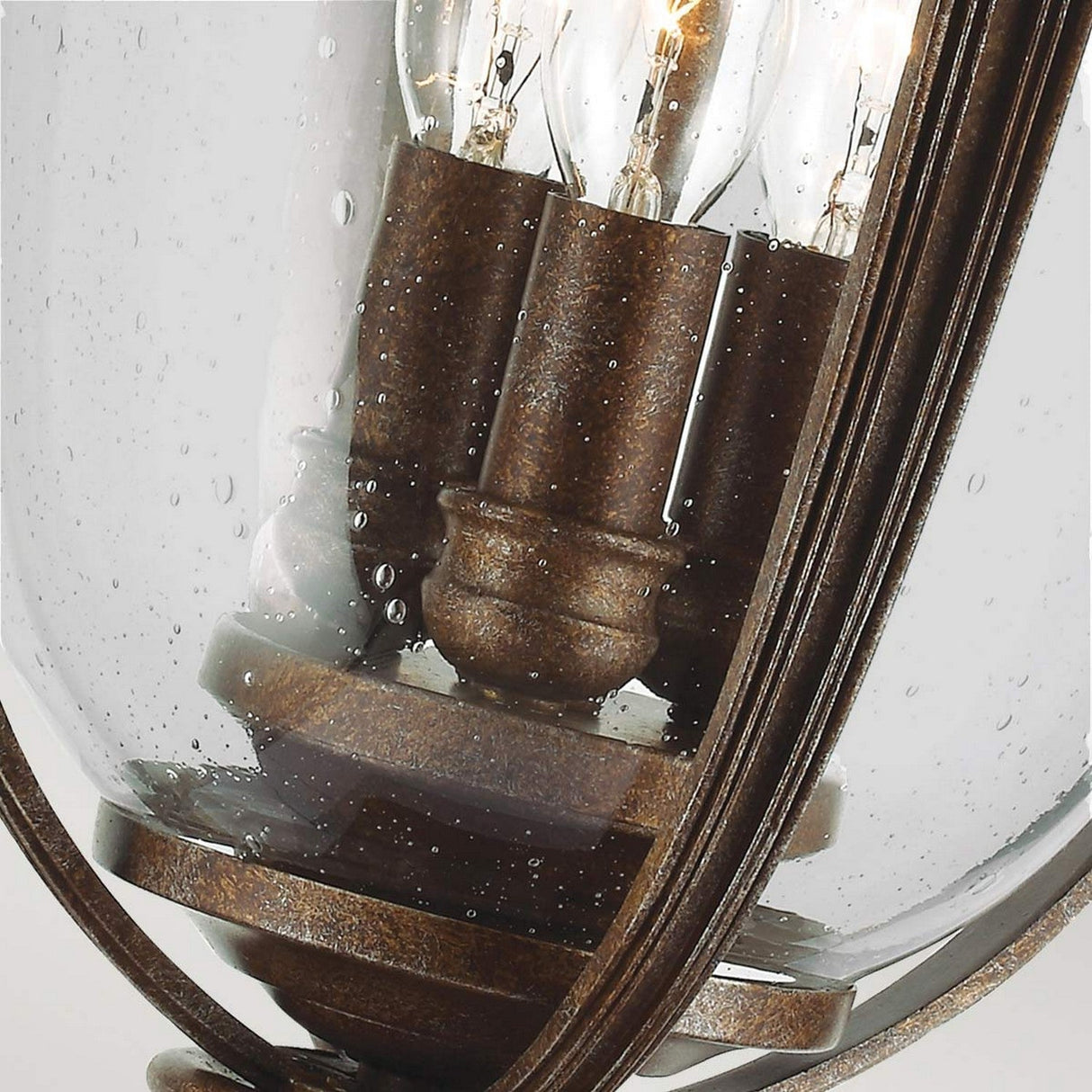 A close-up look at the English Bridle 3 Light Outdoor Large Pedestal in bronze showcases its rustic style and vintage appeal. The pedestal light features elegant metal accents and gracefully curved glass panes. Inside, you can see multiple glowing bulbs, their light enhanced by droplets of water on the glass, which adds texture to its bronze finish.