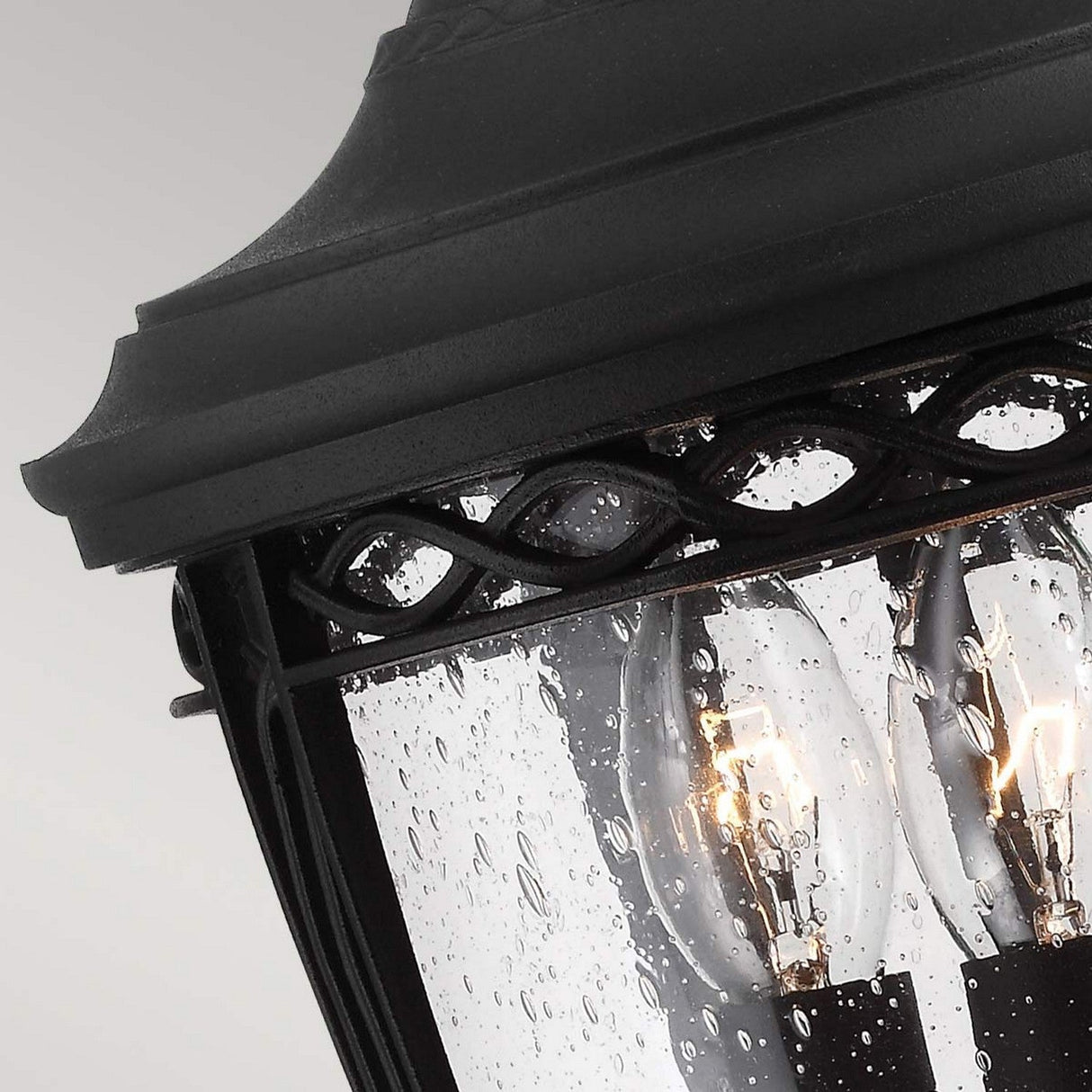 A close-up of the English Bridle 3 Light Outdoor Medium Pedestal in black, featuring two incandescent bulbs illuminating behind clear seeded glass. Raindrops add a textured effect to the glass against a soft gray background.