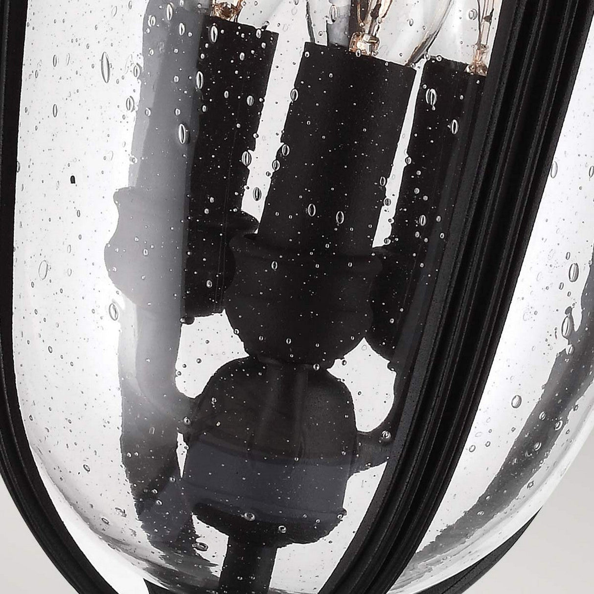 A close-up of the English Bridle 3 Light Outdoor Medium Pedestal - Black highlights its sleek black finish on the metal frame. The clear seeded glass, speckled with water droplets, adds a textured charm while encasing several luminous bulbs.