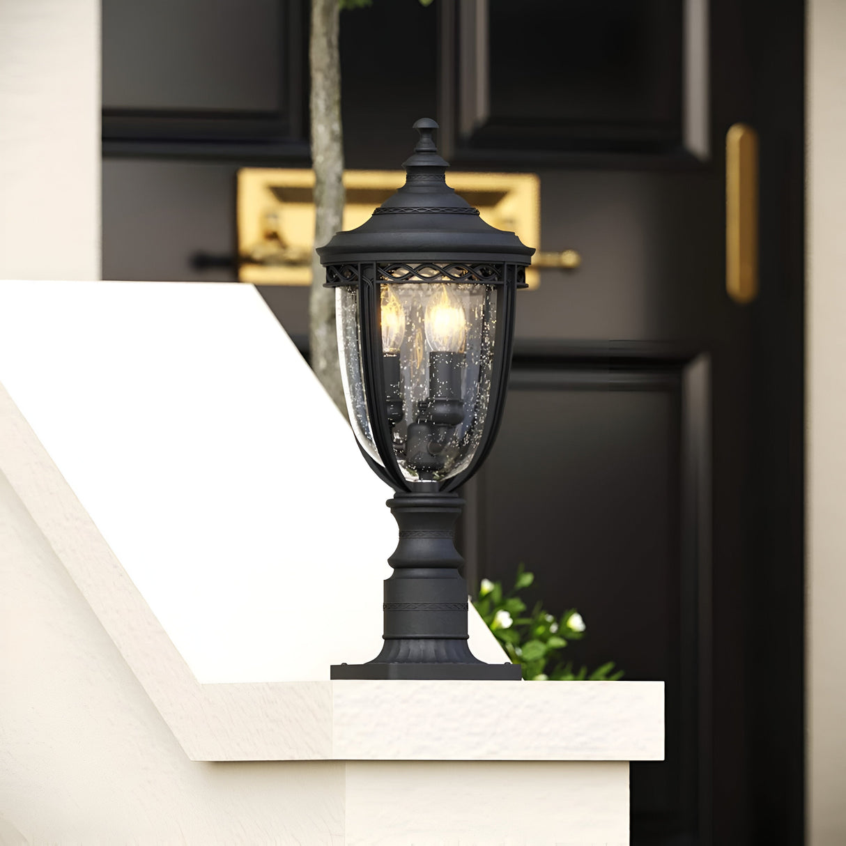The English Bridle 3 Light Outdoor Medium Pedestal in black, featuring ornate detailing and a black finish, is mounted on a white stone pillar. Illuminated with two bulbs and showcasing clear seeded glass, it is set against a dark door with a gold mailbox slot, surrounded by green shrubbery.