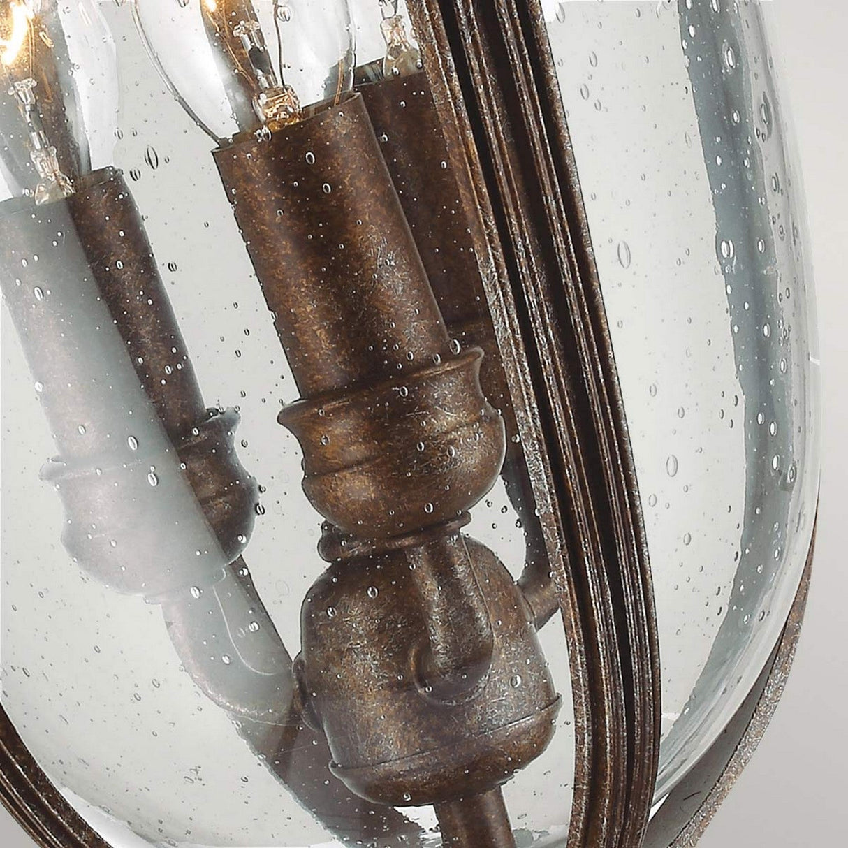 A close-up of the English Bridle 3 Light Outdoor Medium Pedestal - Bronze highlights its vintage charm with raindrop-patterned seeded glass. This bronze outdoor lantern, designed in an English Bridle style, contains multiple candle-shaped bulbs that impart a timeless elegance to any setting.