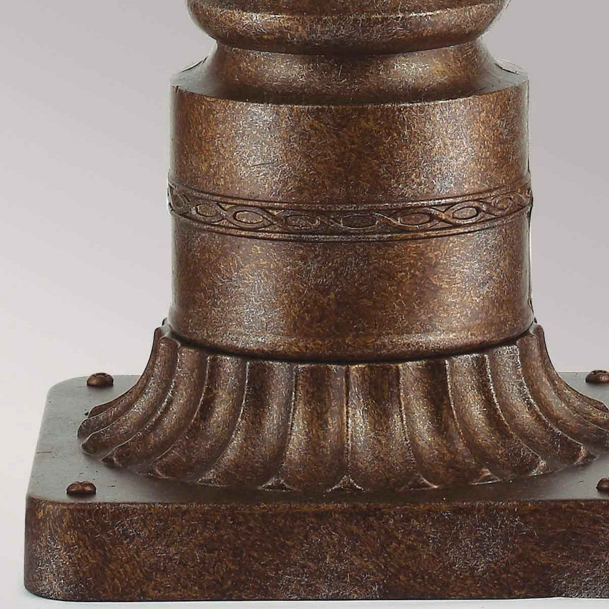 Close-up of a brown, ornate column base with a decorative, carved ring and fluted design resting on a square platform. The antique finish harmonizes beautifully with the English Bridle 3 Light Outdoor Medium Pedestal in bronze nearby, enhancing the elegant ambiance.