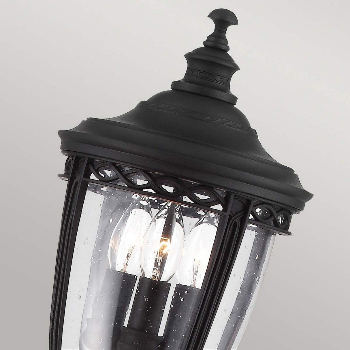 Close-up of the English Bridle 3 Light Outdoor Medium Pillar in black, featuring a decorative top and clear seeded glass panels, showcasing three illuminated candle-shaped bulbs inside. The background is a soft, neutral gray.