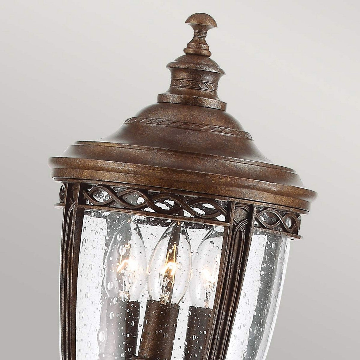 A detailed view of the English Bridle 3 Light Outdoor Medium Pillar Lantern in a bronze finish showcases its ornate design. The clear seeded glass panels exhibit a bubbled texture that reveals three candle-shaped bulbs inside. Featuring intricate detailing, this lantern is adorned with a domed top and a decorative finial.