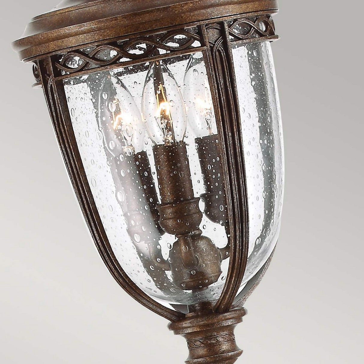 The English Bridle 3 Light Outdoor Medium Pillar Lantern in bronze boasts a vintage design with three candle-shaped lights housed inside a clear seeded glass enclosure. Its detailed metalwork and rustic charm lend an elegant touch.