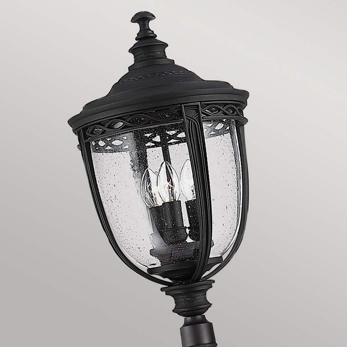 The English Bridle 3 Light Large Lamp Post in black features a traditional design with detailed intricacies, a clear glass encasement, and three candle-like light bulbs. It is set against a plain, light gray background for elegant outdoor lighting.