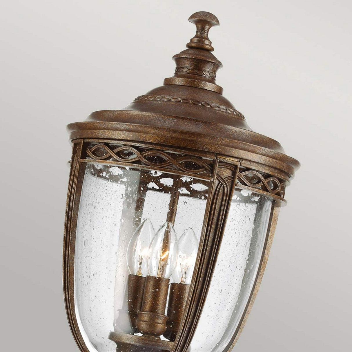 The English Bridle 3 Light Large Lamp Post in a British Bronze finish features a vintage-style design with intricate detailing. It boasts a domed top and clear seeded glass panels, showcasing three candle-like bulbs within. This fixture is displayed at an angle against a neutral background.