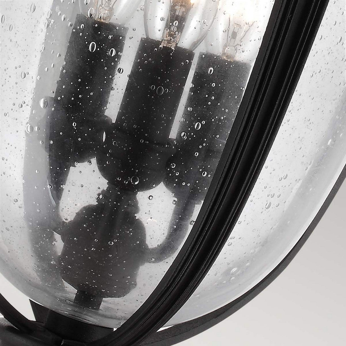 A close-up view of the English Bridle 3 Light Outdoor Large Chain Lantern - Black, featuring its rain-soaked, clear seeded glass with droplets on the surface and three visible light bulbs inside. The black metal frame of the lantern enhances the outdoor lighting ambiance against the clear glass.