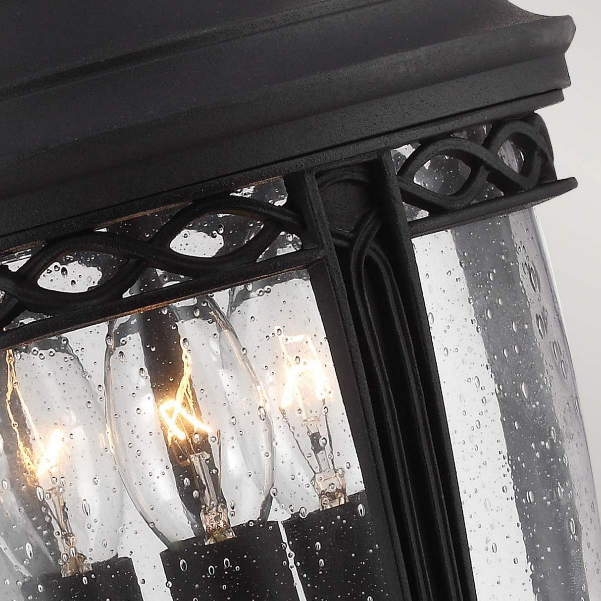A close-up of the English Bridle 3 Light Outdoor Medium Chain Lantern - Black showcases its raindrop-covered glass. Inside, three candle-shaped bulbs emit a warm glow, enhancing the cozy ambiance with vintage charm. The lantern's black metal frame has an intricate woven design at the top, making it ideal for outdoor lighting.