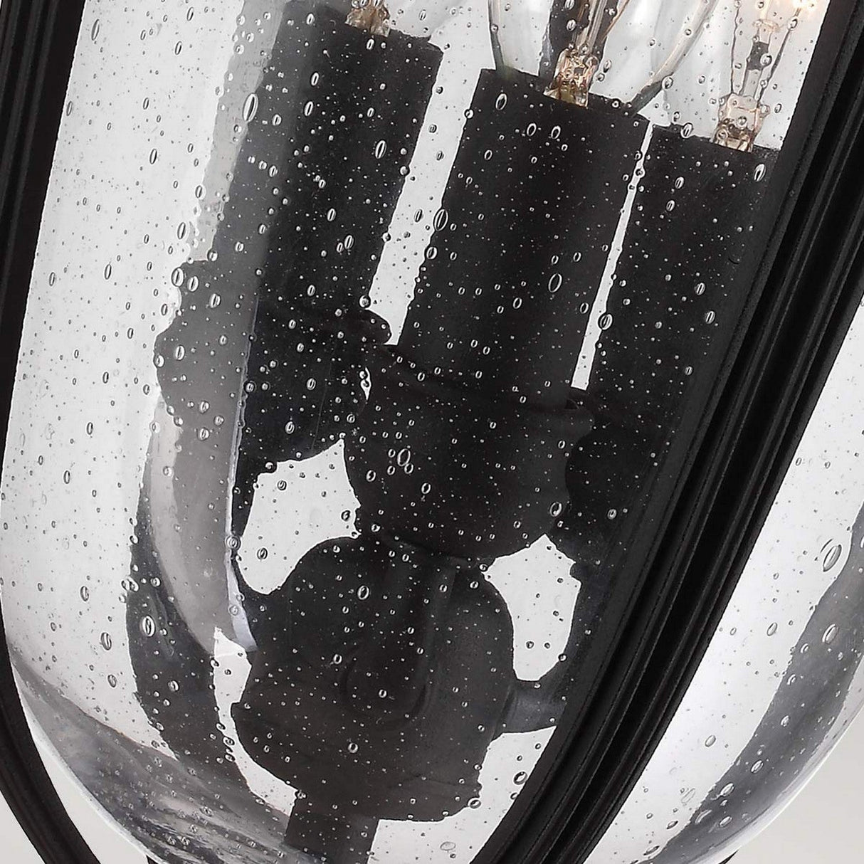 A close-up of the English Bridle 3 Light Outdoor Medium Chain Lantern in Black showcases its clear glass design with visible raindrops on the surface. The interior contains several bulbs mounted on black bases, offering a modern and minimalist look with a hint of vintage style.
