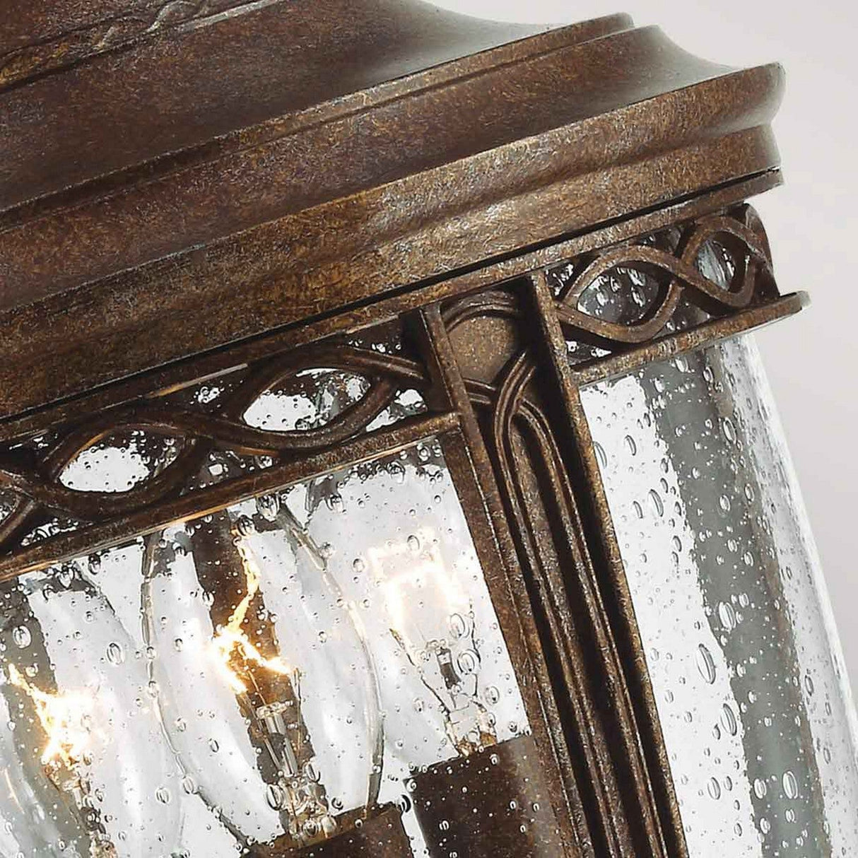 The English Bridle 3 Light Outdoor Medium Chain Lantern in bronze hangs elegantly outside, with its glass speckled by raindrops and softly glowing bulbs that cast a warm light. Its design is accentuated with decorative metalwork along the top.