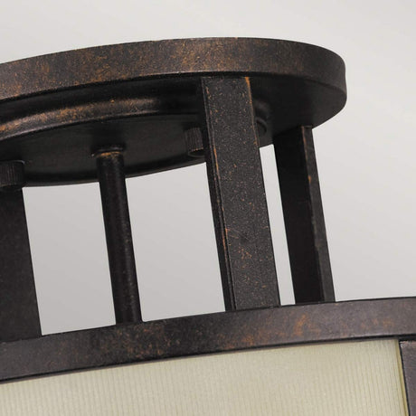 Close-up of the Fusion 3 Light Semi Flush in Grecian Bronze, featuring a cylindrical design with parallel vertical bars and a frosted glass cover. Enhanced by amber ribbed glass shades, this fixture exudes a rustic and industrial look against a pale background.