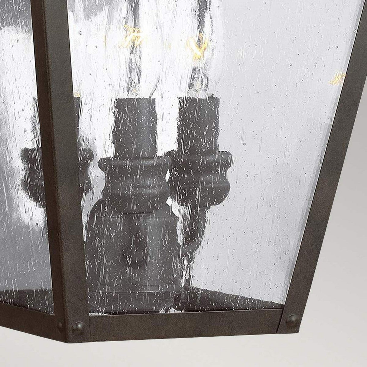 A close-up of the Galena 3 Light Outdoor Large Wall Lantern reveals its rustic candle-style design featuring three bulbs. Its weathered metal frame and textured glass panels contribute to a vintage and elegant look, with bulbs crafted to resemble candles.