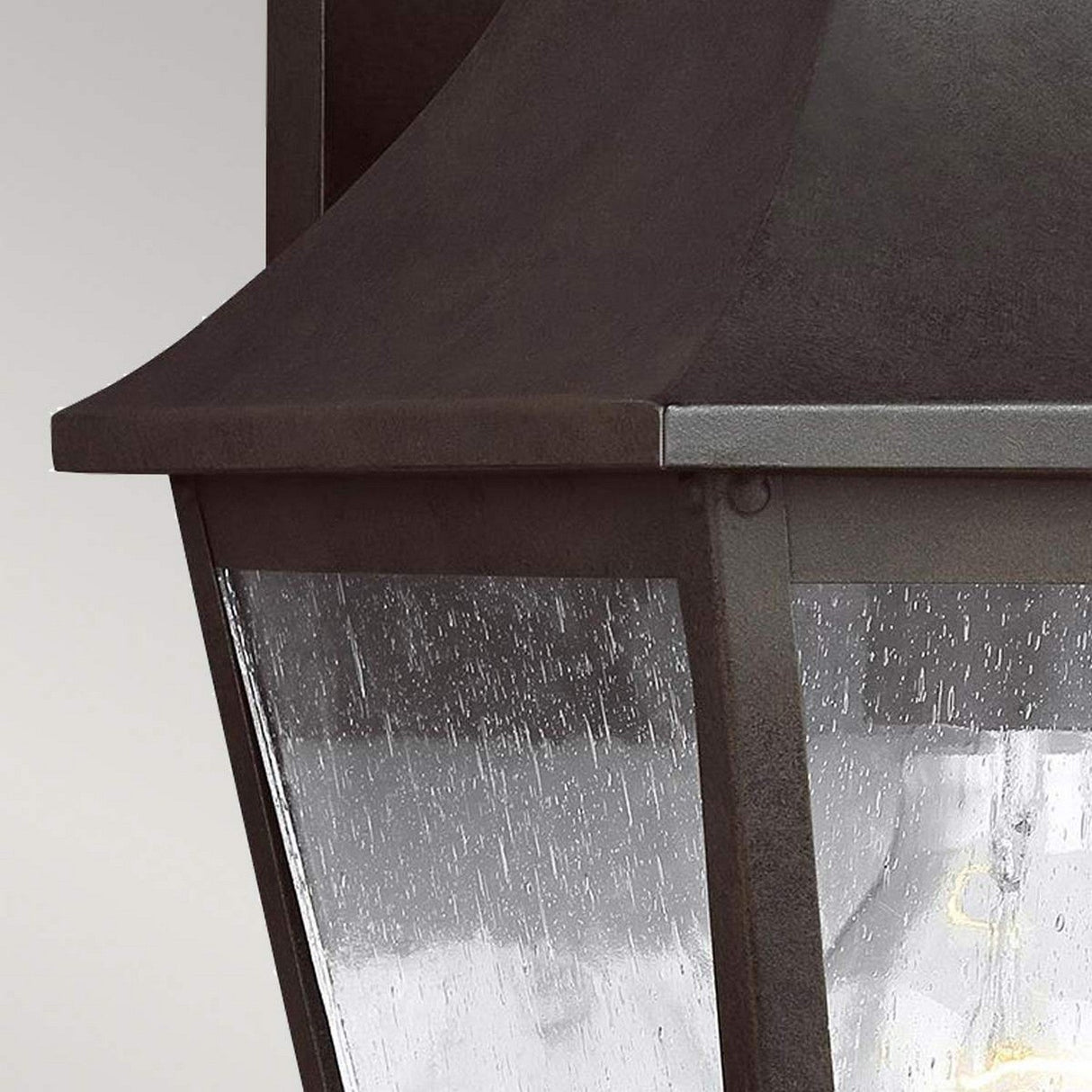 Close-up of the Galena 1 Light Outdoor Medium Wall Lantern in a vintage style, featuring a sable-colored dark metal frame and seeded glass panels. The glass is adorned with rain droplets, partially illuminating the lantern to cast a warm glow.
