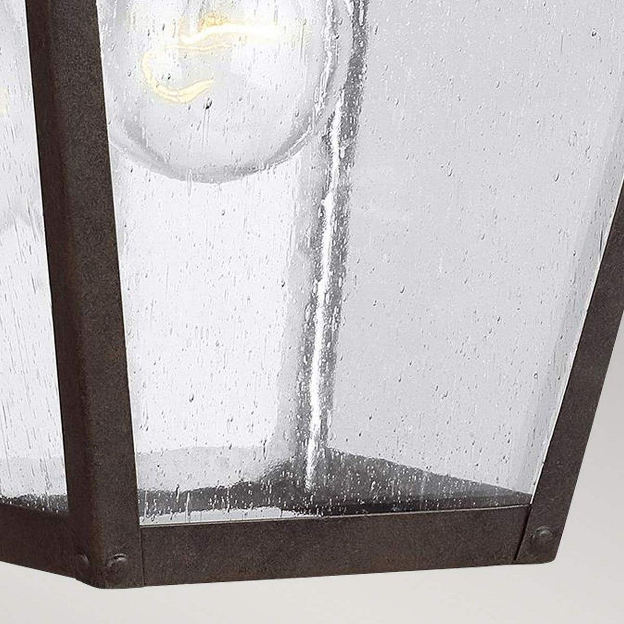 A close-up view of the Galena 1 Light Outdoor Medium Wall Lantern - Sable reveals a glass pane with a raindrop texture, encased in a metal frame similar to seeded glass. Inside, the clear incandescent bulb is partially visible, emitting a soft glow. The surface of the pane captures the light beautifully, creating a shimmering effect akin to an IP44-rated outdoor lantern.