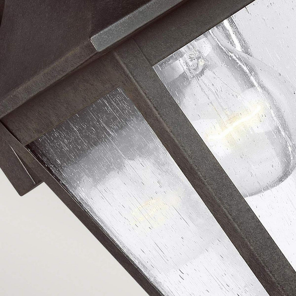 A close-up of the Galena 1 Light Outdoor Small Wall Lantern in grey showcases a visible light bulb inside. The seeded glass creates a rainy effect, and its dark metal frame is stylishly set against a neutral backdrop. With an IP44 rating, this lantern is designed for durability against the elements while enhancing any setting with its charm.