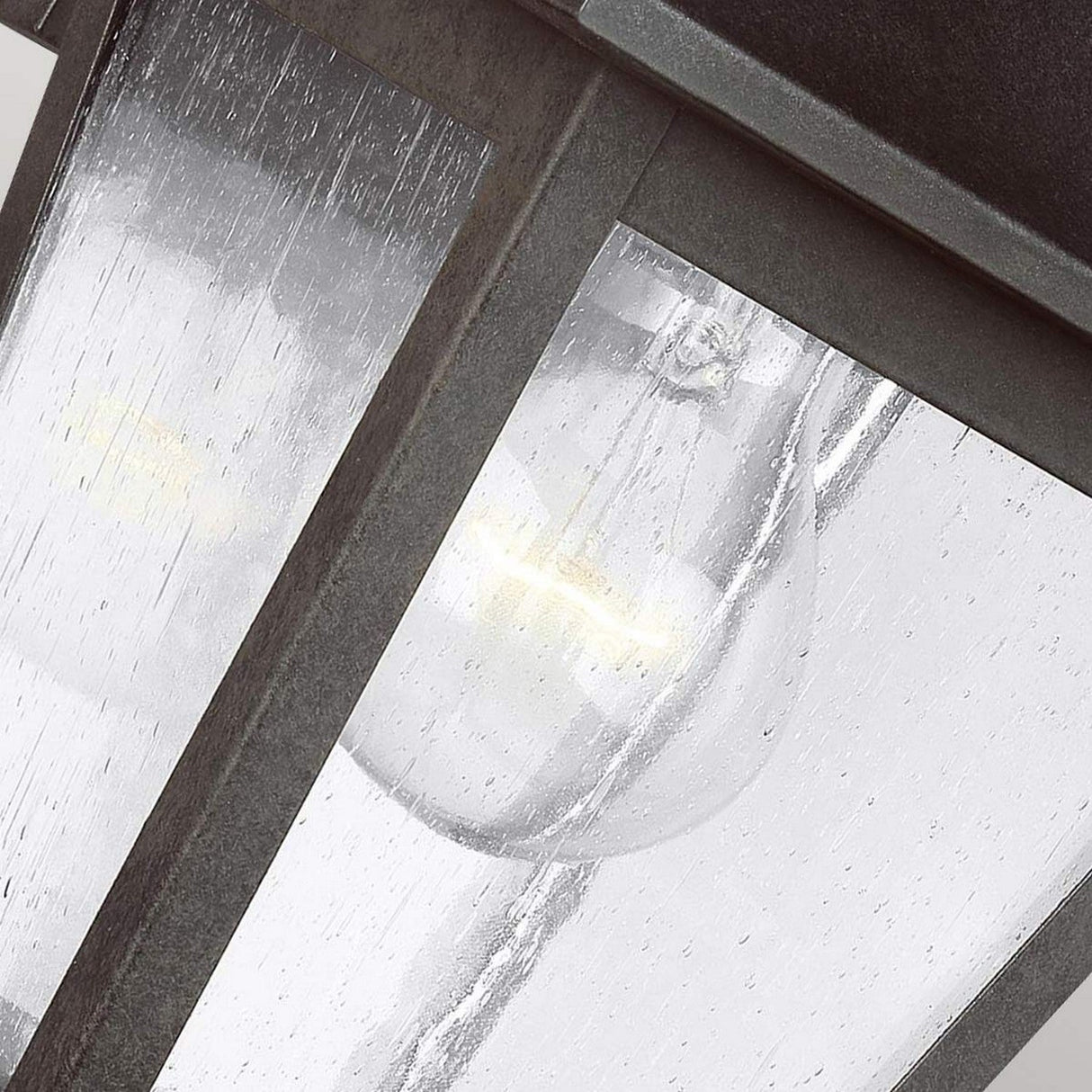 A close-up of the Galena 1 Light Outdoor Small Wall Lantern - Grey reveals a glowing bulb inside. The seeded glass panels are speckled with raindrops, complementing the metal frame's weathered texture. Its soft lighting creates a warm and inviting atmosphere, and its IP44 rating ensures durability against the elements.