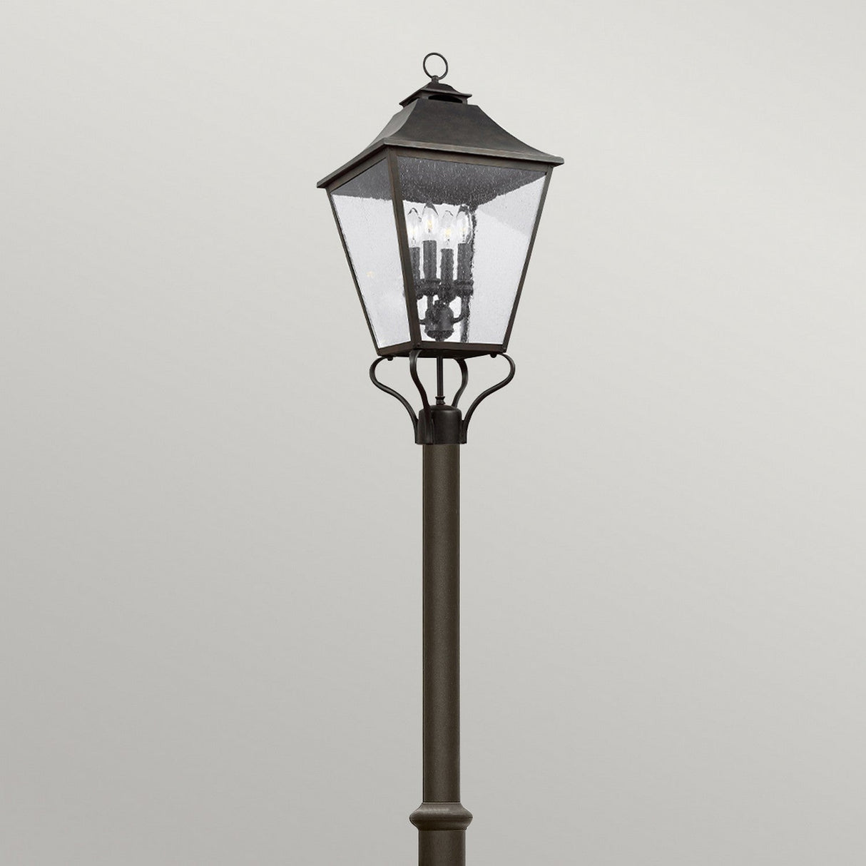 The Galena 4 Light Lamp Post - Grey features a classic outdoor design with a black metal frame and clear seeded glass panels, showcasing multiple light bulbs inside. Set against a plain, light gray background, it enhances the allure of outdoor lighting in any setting.