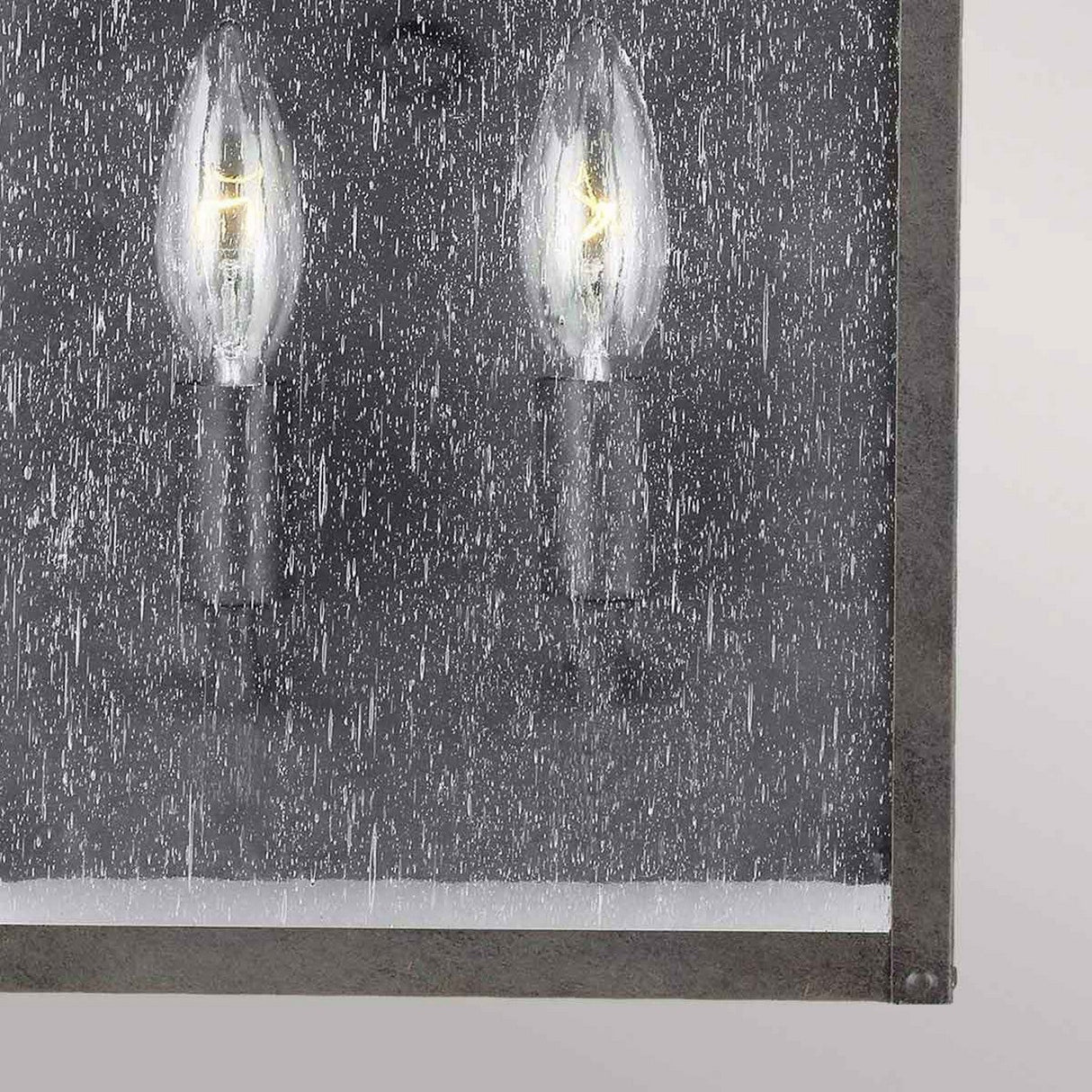 The Galena 2 Light Outdoor Half Lantern in Grey features a close-up view of its two candle-like bulbs behind a textured, seeded glass pane, creating a rain-speckled effect. Its metal frame adds an industrial look that contrasts beautifully with the soft glow of this exterior lighting piece.
