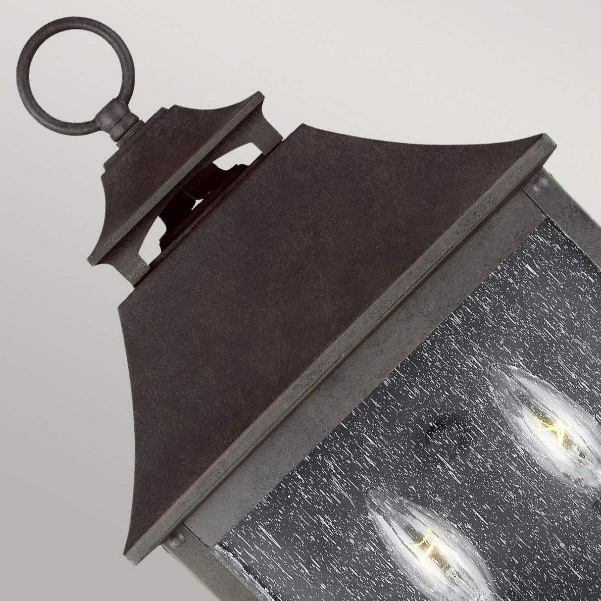 A close-up of the Galena 2 Light Outdoor Half Lantern in dark metal with a rustic finish. This exterior lighting fixture showcases two candle-shaped light bulbs behind seeded glass, which has a textured, frosted appearance, against a plain light gray background.