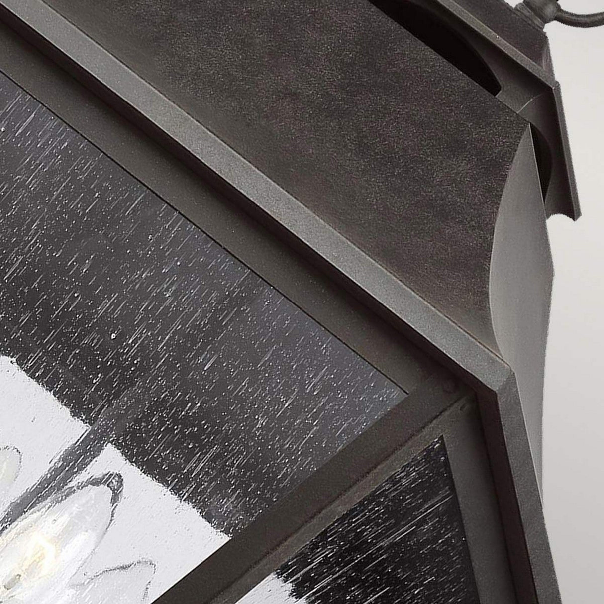 Close-up of the Galena 4 Light Outdoor Chain Lantern in grey, featuring seeded glass panels that partially reflect light. The metal frame showcases a weathered texture with a visible light bulb inside. The angle reveals only part of the lantern's geometric design, indicative of its IP44 rated durability.