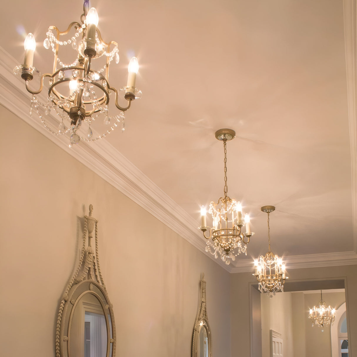 An elegant hallway features three ornate chandeliers casting a warm glow on the beige walls. Two Gianna 4 Light Chandeliers in Gilded Silver, adorned with hand-cut crystal embellishments and showcasing Italian design, enhance the space's classic ambiance while decorative mirrors reflect their captivating light.