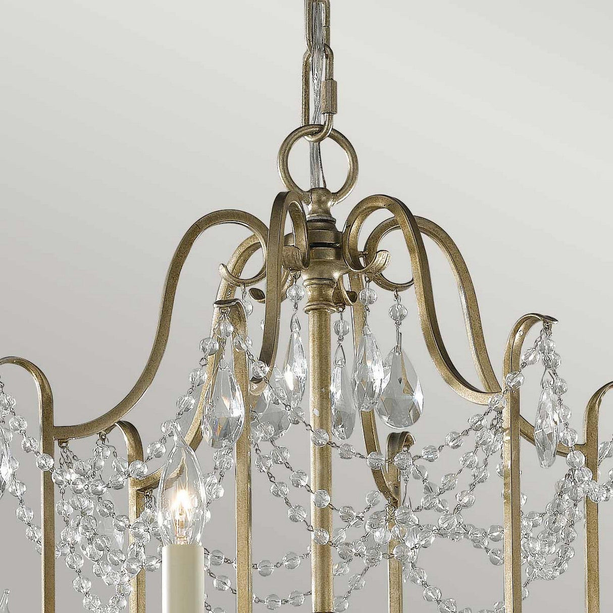 A close-up of the Gianna 6 Light Chandelier - Gilded Silver reveals its intricate design featuring gold-colored metal arches adorned with strings of hand-cut crystals. The chandelier's finish beautifully complements the small candle-shaped bulbs, enhancing its elegance against a simple backdrop.