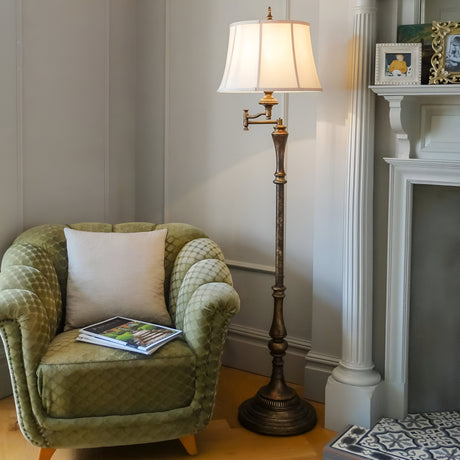 A cozy living room corner with a green quilted armchair, a white pillow, a book on the chair, and the dark brown Gibson 1 Light Floor Lamp adorned with a Desert Linen Fabric Shade. The room features light gray walls, a white fireplace with decorative molding, and a photo frame on the mantel.