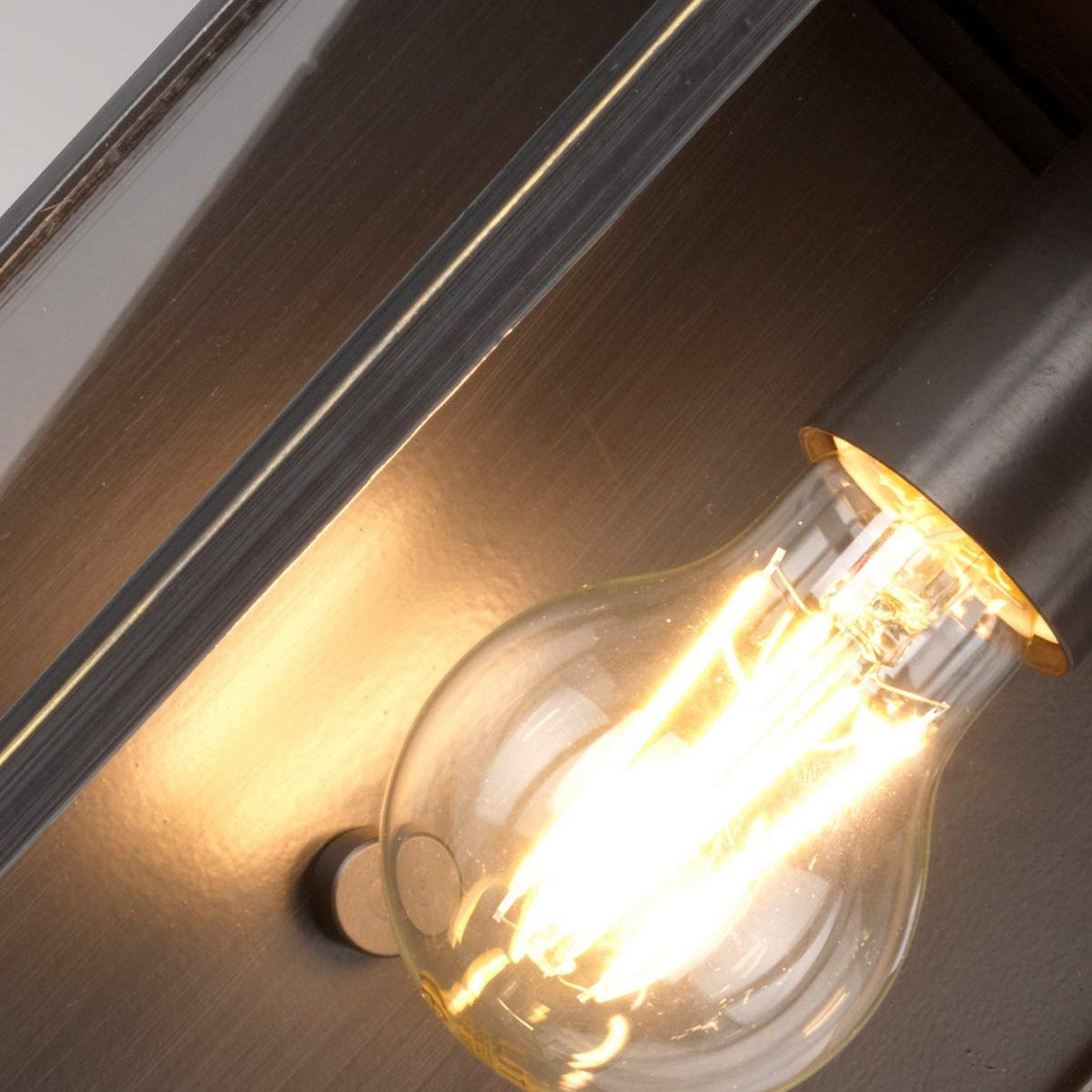 A lit Glenview 1 Light Outdoor Medium Wall Lantern in antique bronze is mounted on a wall. The close-up image emphasizes the glowing filament inside the glass bulb, casting a warm light and creating a cozy atmosphere reminiscent of a prairie-style design.