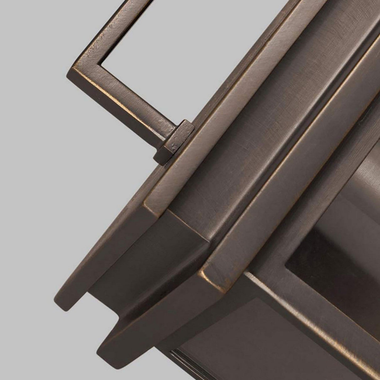 Close-up of the Glenview 1 Light Outdoor Small Wall Lantern in an Antique Bronze finish, showcasing parallel lines and angular elements against a plain gray background. The image emphasizes the metallic texture and geometric form of the lantern.