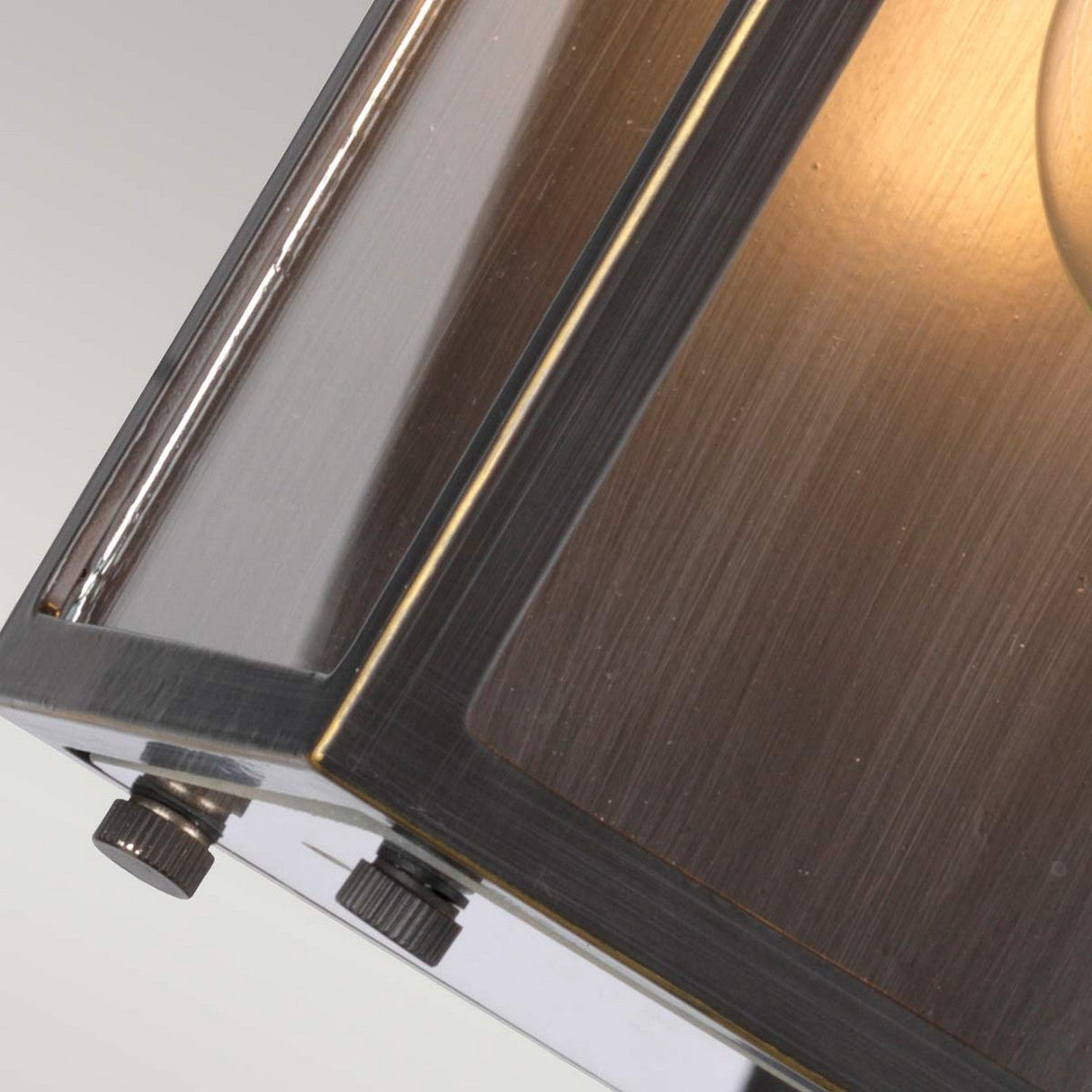 Close-up of the metallic corner of the Glenview 1 Light Outdoor Small Wall Lantern in an Antique Bronze finish, highlighting its brushed metal texture and reflective surfaces. The lantern is illuminated, casting a warm glow with visible screws on its edges.