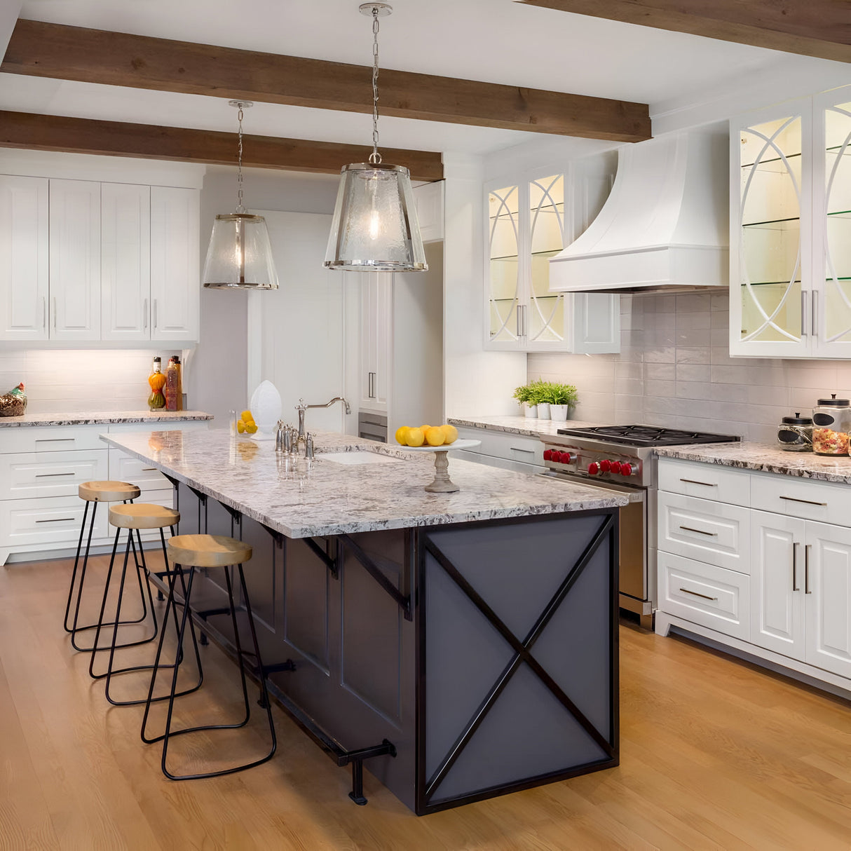 A modern kitchen with a large marble island features three wooden bar stools, while Harrow 1 Light Medium Pendant in polished nickel elegantly hangs above. The brightness is enhanced by white cabinets with seeded glass doors, and an array of fruits and greenery adds a splash of color to the space.
