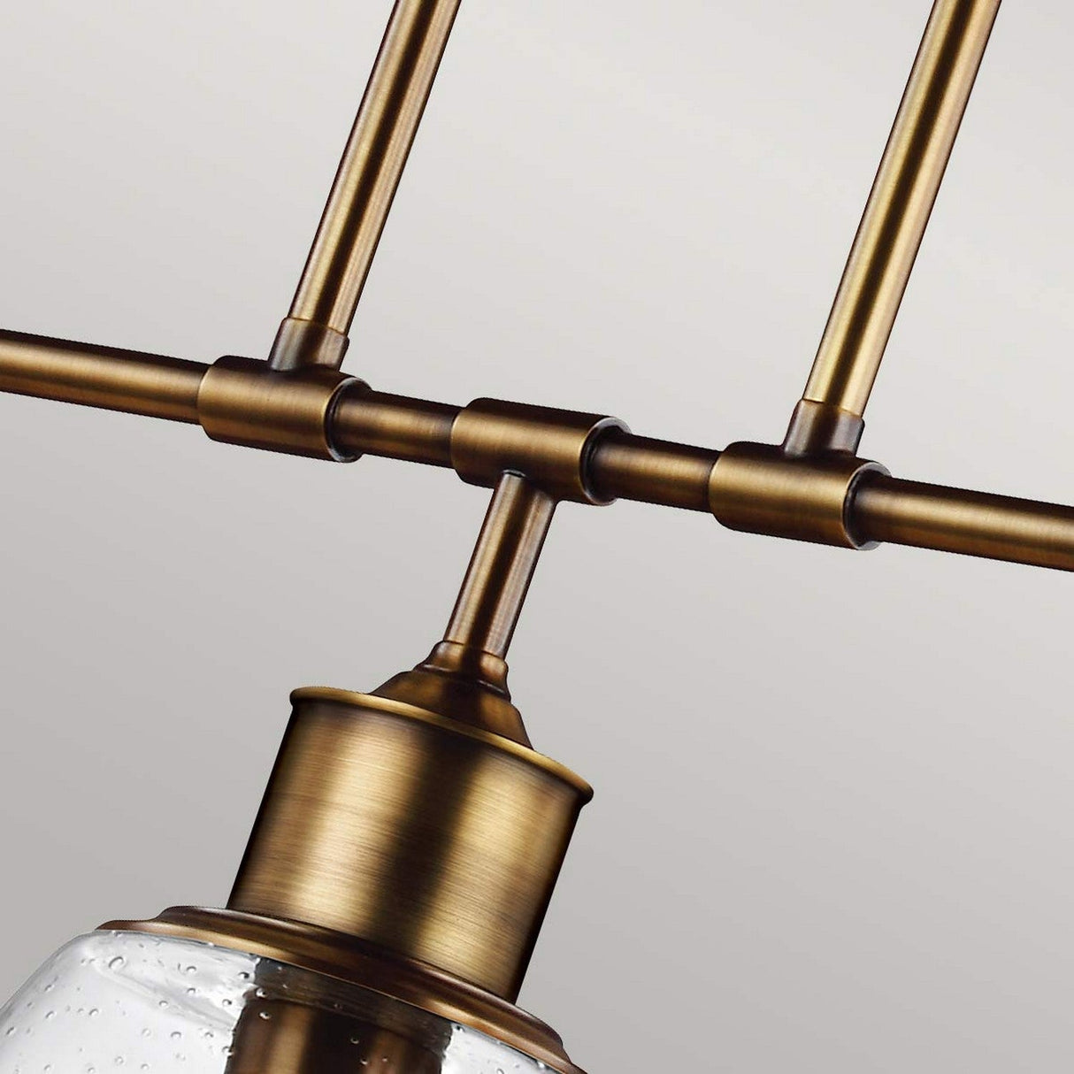 Close-up of the Hobson 3 Light Island Light in aged brass, showcasing its modern design. The fixture features sleek rods and a clear, seeded glass shade with a textured surface, set against a neutral background.
