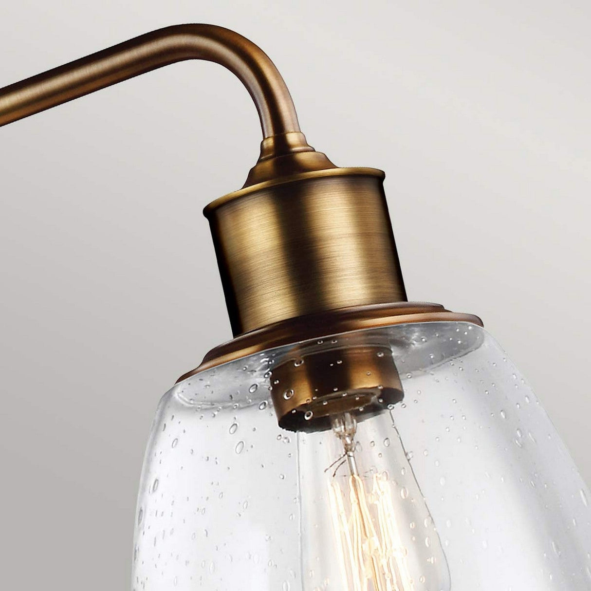 Close-up of the Hobson 3 Light Island Light in aged brass, showcasing its antique style with seeded glass shades. The spotted glass resembles water droplets, revealing an Edison bulb inside that emits a warm glow. The background is a soft gray.