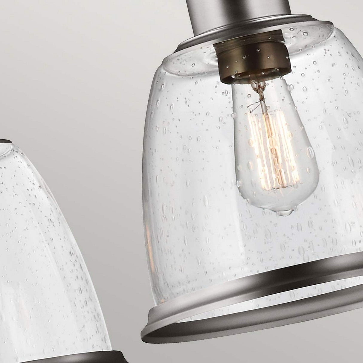 A close-up of the Hobson 3 Light Island Light in satin nickel reveals two clear pendant lights with vintage-style bulbs inside each. The seeded glass is adorned with a subtle bubble texture, enhancing the industrial design against a neutral background.