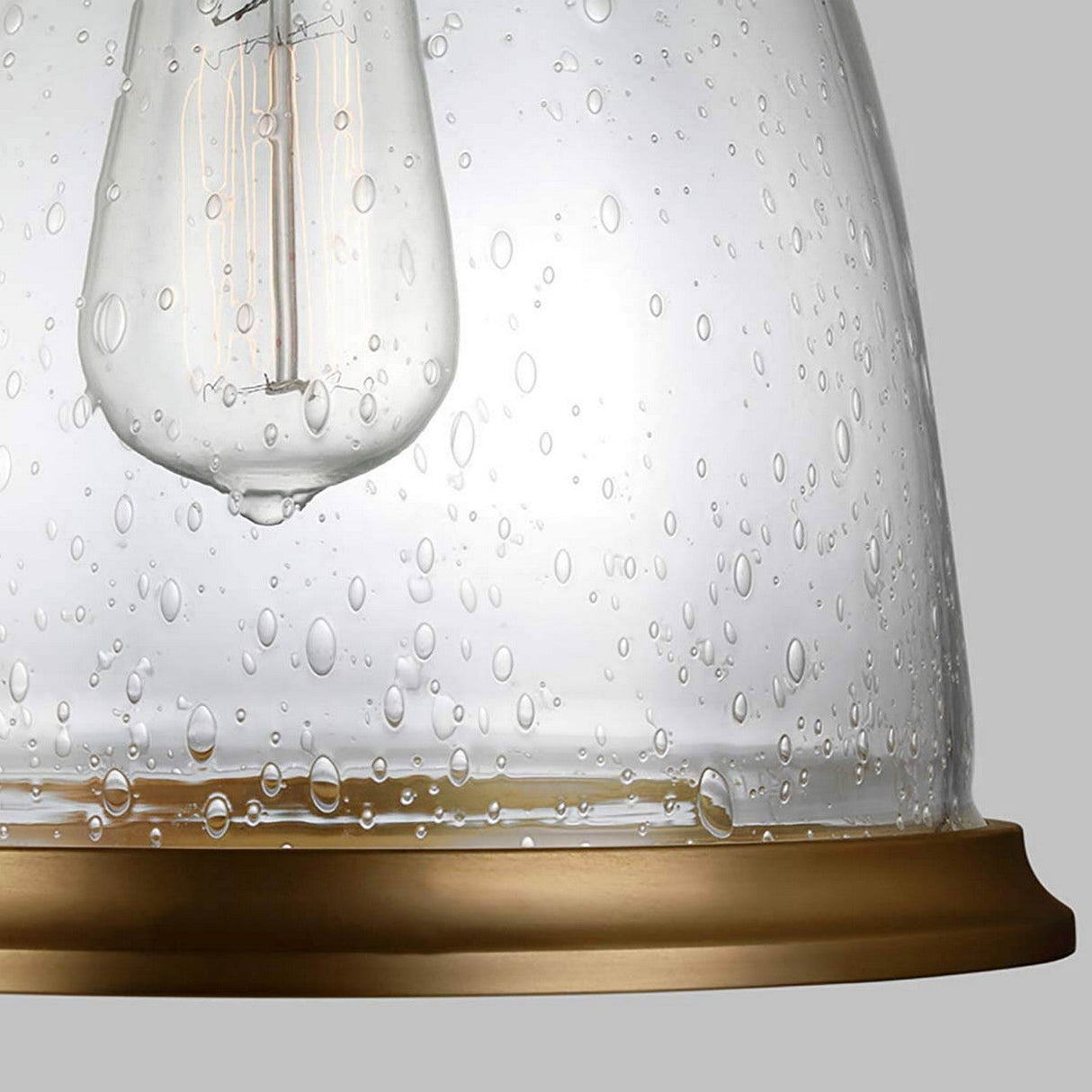 A close-up view of the Hobson 1 Light Medium Pendant - Aged Brass, featuring its seeded glass shade that displays a water droplet texture. The light bulb is attached to a circular base with an aged brass finish, all set against a neutral gray background.