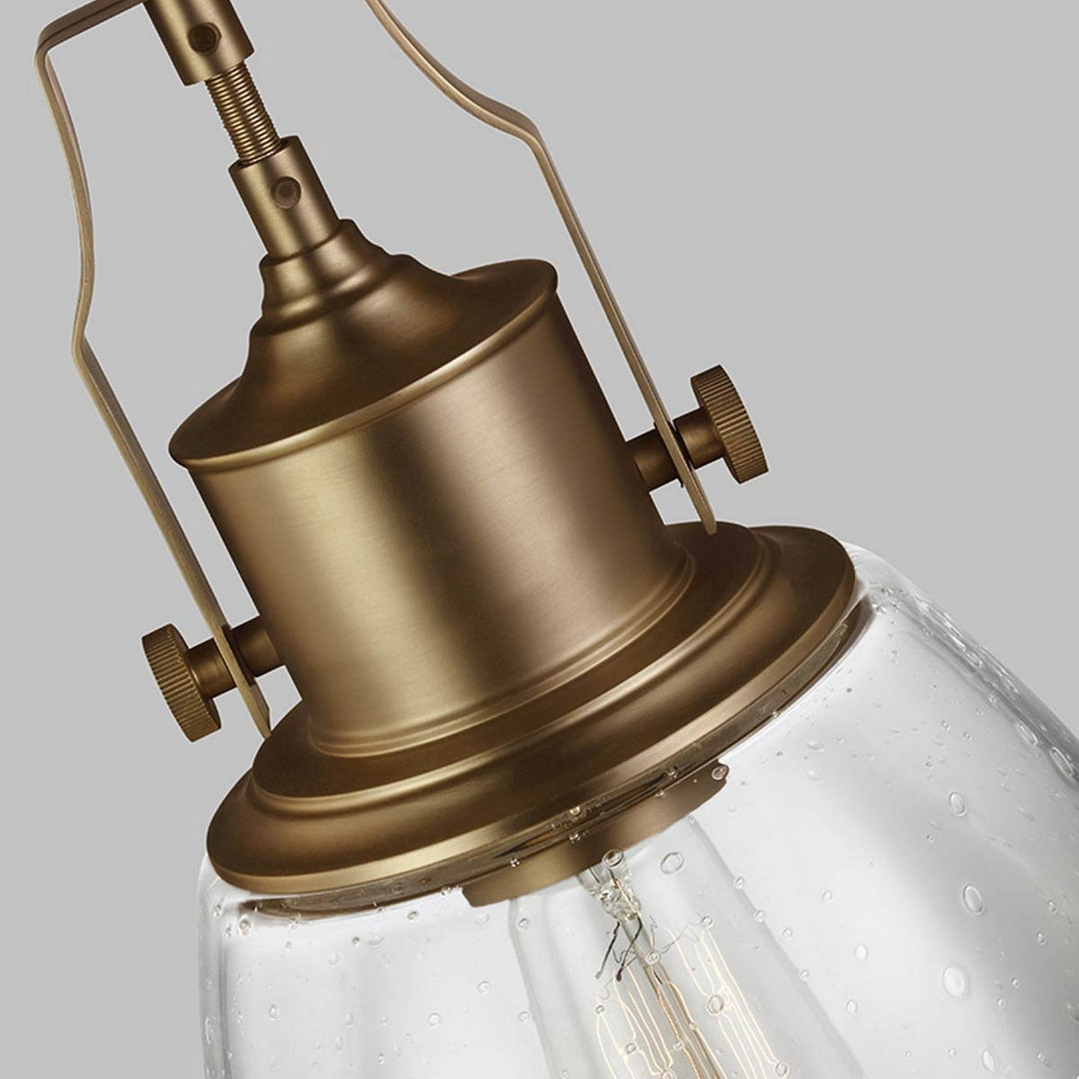 Close-up of the Hobson 1 Light Medium Pendant - Aged Brass, featuring an industrial-style design with an aged brass finish on the fixture and a clear seeded glass shade adorned with small air bubbles. The visible bulb adds charm against the neutral gray background.