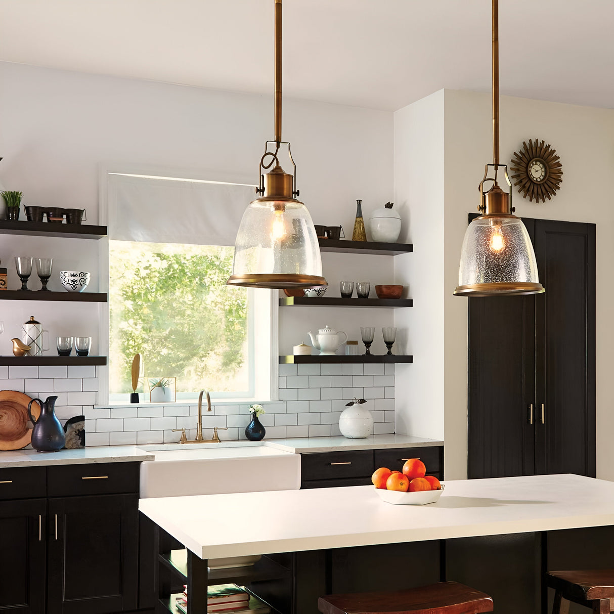 A modern kitchen features dark cabinetry, a white subway tile backsplash, and golden accents. Two Hobson 1 Light Medium Pendants in Aged Brass with seeded glass shades are installed above a white island counter adorned with a bowl of oranges. Open shelves showcase dishes and decor, while a large window provides natural light and a view of greenery.