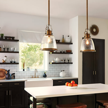 A modern kitchen features dark cabinetry, a white subway tile backsplash, and golden accents. Two Hobson 1 Light Medium Pendants in Aged Brass with seeded glass shades are installed above a white island counter adorned with a bowl of oranges. Open shelves showcase dishes and decor, while a large window provides natural light and a view of greenery.