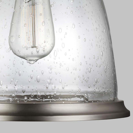 A detailed view of the Hobson 1 Light Medium Pendant showcases a vintage-style bulb encased within a raindrop-textured seeded glass shade. The design features an industrial flair complemented by a satin nickel finish, allowing the bulb's filaments to be partially visible through the textured glass.